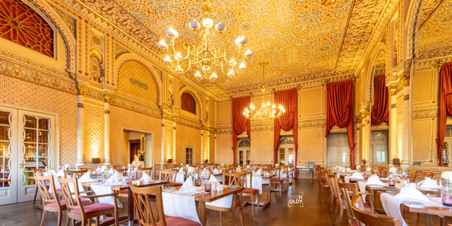 Ein eleganter Speisesaal mit hohen Decken, großen Kronleuchtern, roten Vorhängen und gedeckten Tischen.