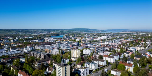 Luftaufnahme einer Stadt mit mehreren Gebäuden, Straßen und Bäumen, im Hintergrund ein Fluss und Hügel.