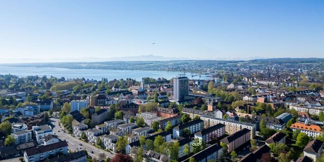 Luftaufnahme einer Stadt mit Gebäuden, Straßen und einem großen See im Hintergrund, umgeben von grünen Bäumen und Hügeln.