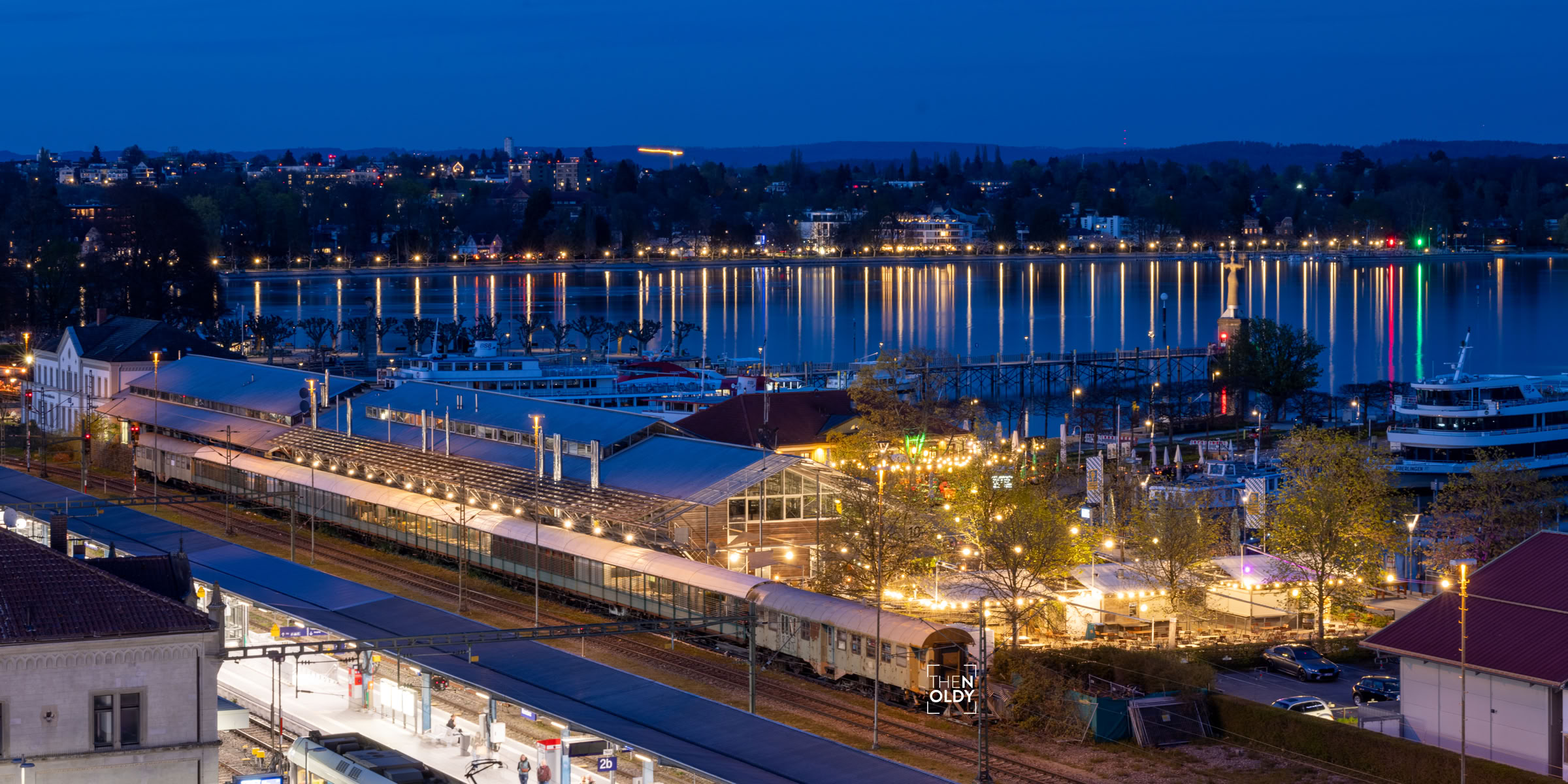 Bahnhof bei Nacht mit beleuchteten Zügen und Gebäuden, im Hintergrund ein Fluss mit Booten und beleuchtete Stadt.