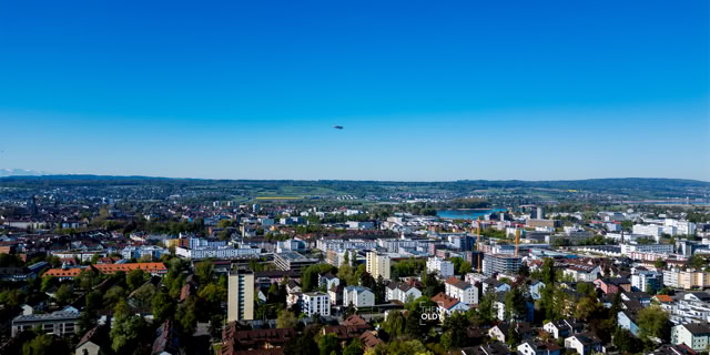 Stadtpanorama mit Gebäuden, Bäumen und einem klaren blauen Himmel im Hintergrund.