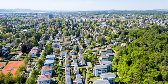 Luftaufnahme einer Wohngegend mit vielen Häusern, Bäumen und einem Tennisplatz, im Hintergrund sind Hügel sichtbar.