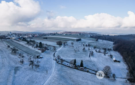 Luftaufnahme einer verschneiten Landschaft mit Feldern, Bäumen und einem kleinen Dorf im Hintergrund unter einem bewölkten Himmel.